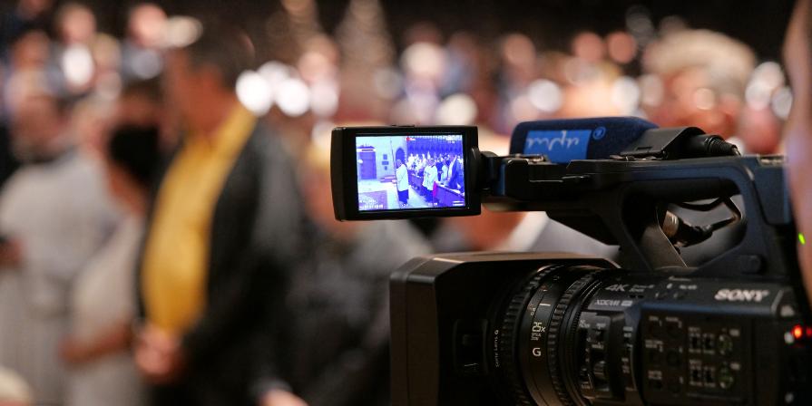 Eine professionelle Videokamera steht im Vordergrund und filmt eine Veranstaltung. Auf dem aufgeklappten Kameradisplay ist eine Gruppe von Menschen in einem Innenraum zu sehen. Im unscharfen Hintergrund befinden sich weitere Personen, die offenbar an der Veranstaltung teilnehmen.