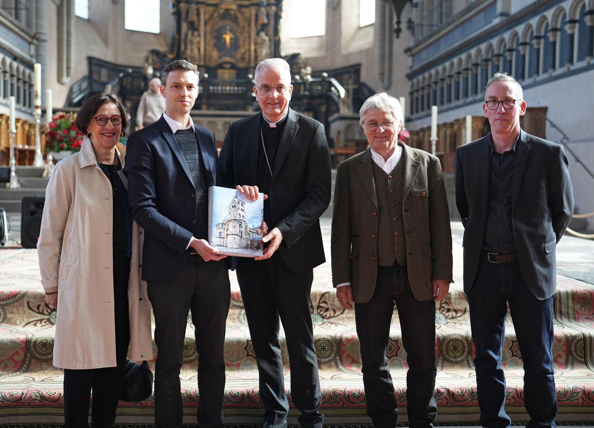Vier Männer und eine Frau stehen im Trierer Dom vor dem Altar. Zwei Männer halten ein Buch in der Hand.